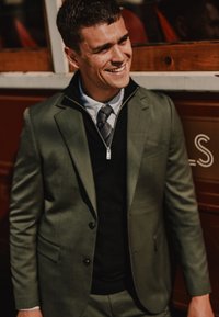 Alyvuogių žalios spalvos švarkas su siauru siluetu, uždėtas ant juodos megztinio. Yra užtrauktukas priekyje ir pilkų bei baltų dryžuotų marškinių.