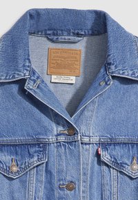 Svetlo modra denim jakna s klasičnim ovratnikom, dvema žepoma na prsih in bronastimi gumbi. V notranjosti ovratnika je usnjen etiketa z blagovnimi podrobnostmi.