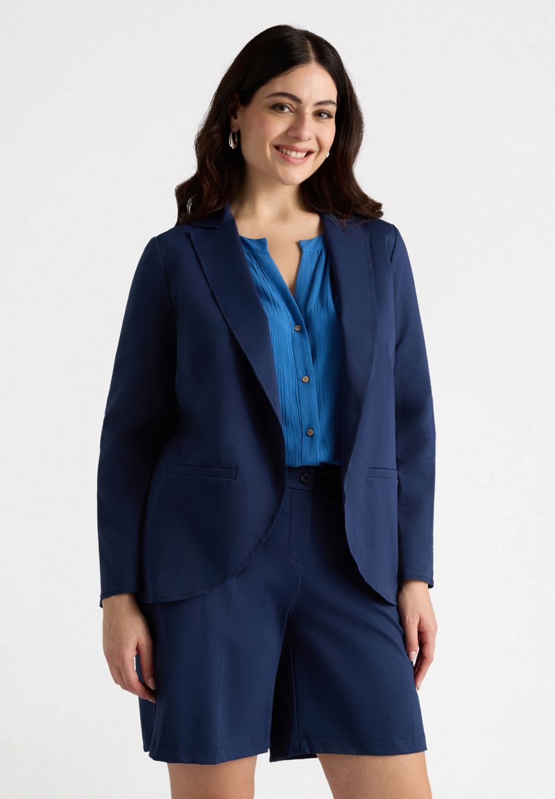 Navy blazer dan celana pendek serasi dipadukan dengan blus biru berlipat yang memiliki kancing. Blazer dilengkapi dengan kerah dan saku, serta memiliki tekstur kain yang halus.