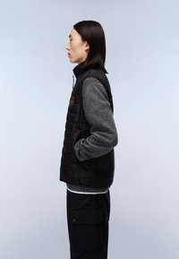 Svart vadderad väst med grå stickade ärmar. Har en hög krage och logotypdetalj. Bärs över en grå tröja, ihop med mörka cargobyxor.