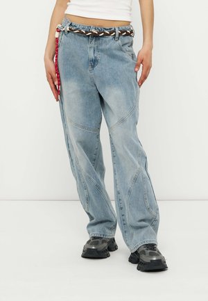 Hellblaue Jeans mit lockerem Sitz und geschwungenen Nahtdetails, brauner geflochtener Gürtel und klobige grau-schwarze Turnschuhe an einer Person, die ein weißes, kurz geschnittenes Oberteil trägt.