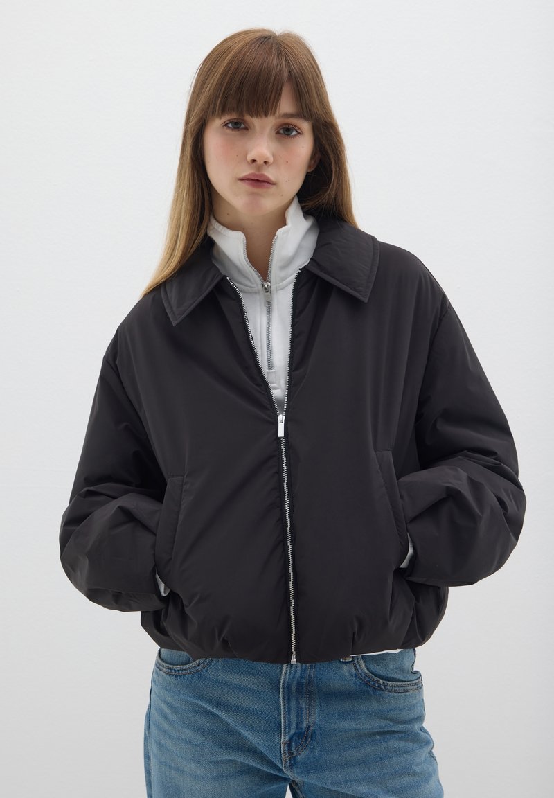 Schwarze Bomberjacke mit Kragen, Reißverschluss vorne und seitlichen Taschen, übereinander getragen mit einem weißen Reißverschluss-Oberteil, kombiniert mit hellblauen Denim-Jeans.