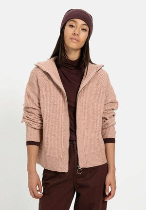 Lyserød ribstrikket cardigan med lynlås og oversized krave. Båret over en mørkerød turtleneck, suppleret med en matchende beanie.