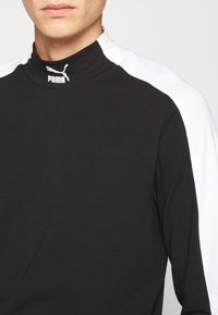 Czarna bluzka z długim rękawem i wysokim kołnierzem. Posiada kontrastujące białe panele na ramionach oraz małe białe logo Puma na kołnierzu. Miękka tekstura materiału.