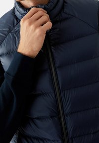 Granatowa pikowana kamizelka z pikowanymi panelami, wysokim kołnierzem i centralnym zamkiem błyskawicznym. Miękka, błyszcząca faktura z eleganckim, smukłym krojem.