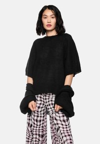 Schwarzer, übergroßer Pullover aus strukturiertem Stoff mit weiten Ärmeln, kombiniert mit gemusterten Karohosen in Schwarz, Weiß und Pink.