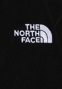 Fekete polár anyag, amelyen a "THE NORTH FACE" fehér logója látható, félkör alakú dizájnnal. Puha tapintású, minimalista stílusú.