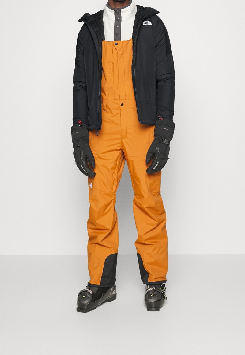 Personne portant un pantalon de ski orange à bretelles, une veste d'hiver noire, des gants noirs et des chaussures de ski, debout devant un fond uni.