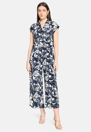 Femme debout portant une combinaison sans manches imprimée de fleurs bleu marine et blanches, à jambes larges, avec des sandales à talons beiges.