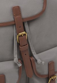 Grå tygryggsäck med bruna läderdetaljer, ett lock med en mässingsspänne och dragsnöre för säker förvaring.