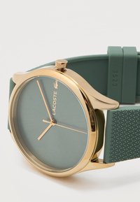 Ρολόι χειρός Lacoste με χρυσό τόνο, απλό πράσινο καντράν, χρυσά δείκτες και υφασμένο πράσινο λουράκι από σιλικόνη.