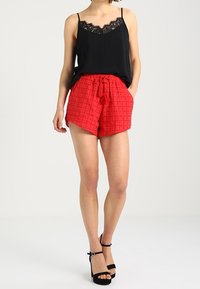 Svart camisole med spetsdetaljer, tillsammans med röda shorts med en strukturerad mönstring, knytmidja och svarta sandaler med höga klackar och remmar.