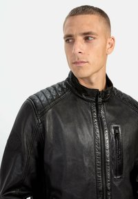Sort læderjakke med høj krave, kviltet skulderdetalje og lynlåslomme på brystet. Glat tekstur med et elegant design.