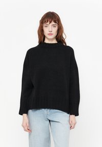 Crni pleteni džemper s visokim ovratnikom, oversized siluetom, spuštenim ramenima i rebrastim manžetama. Nositi s svijetlim plavim trapericama.