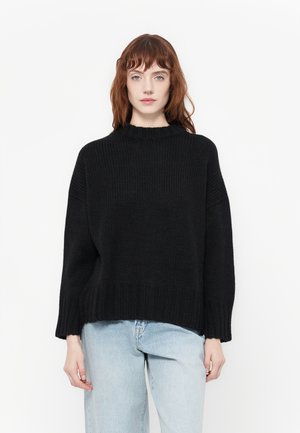 Svart strikket genser med høy hals, oversized silhuett, drop shoulders og ribbestrikkede mansjetter. Båret med lyseblå jeans.
