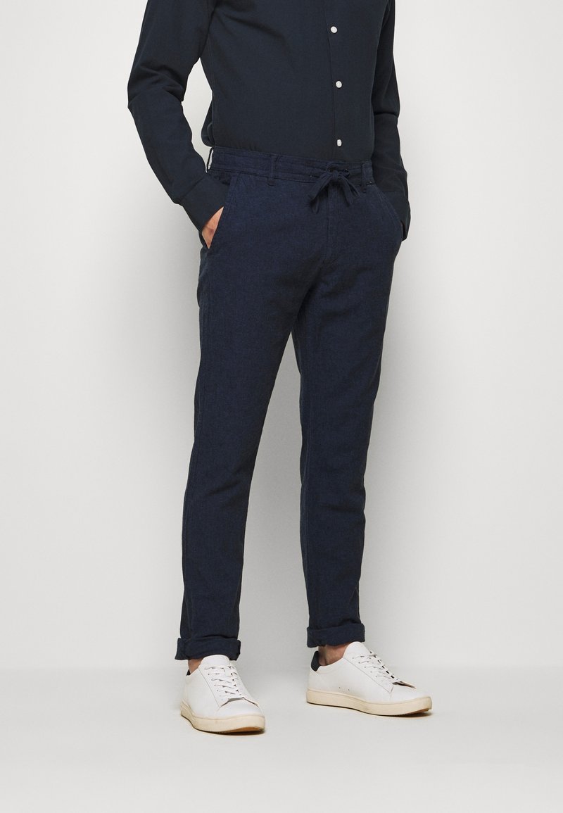 Homme portant un pantalon bleu marine à cordon, une chemise noire boutonnée, des baskets blanches, les mains dans les poches, debout devant un fond uni.