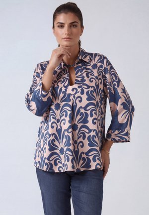 Vrouw met donker haar in een roze en donkerblauwe blouse met lange mouwen met patroon, poserend met haar hand dicht bij de kin tegen een effen achtergrond.