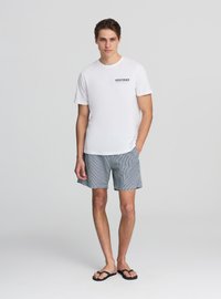 Maglietta bianca a maniche corte con scritta nera "VENICE BEACH", abbinata a bermuda da bagno a righe blu e bianche e infradito nere.