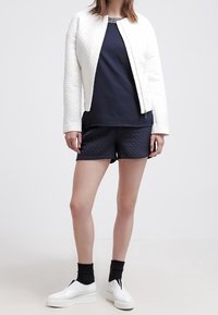 Vit quiltad jacka, marinblå överdel, marinblå quiltade shorts och vita plattformssneakers. Outfiten har en rund halsringning och kontrasterande texturer.