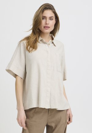 Femeie cu păr lung, castaniu deschis, purtând o cămașă lejeră bej cu mâneci scurte și nasturi și pantaloni maro, stând în picioare pe un fundal alb simplu.