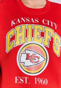 Sudadera roja con la inscripción "KANSAS CITY CHIEFS" en grandes letras amarillas, un logo circular con "KC" y "EST. 1960" en texto blanco. Tejido suave.