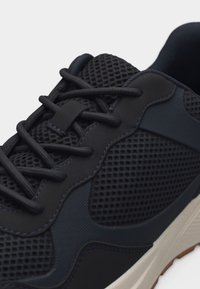 Baskets noires en tissu mesh, avec des superpositions synthétiques, des lacets noirs et une semelle en caoutchouc blanche.