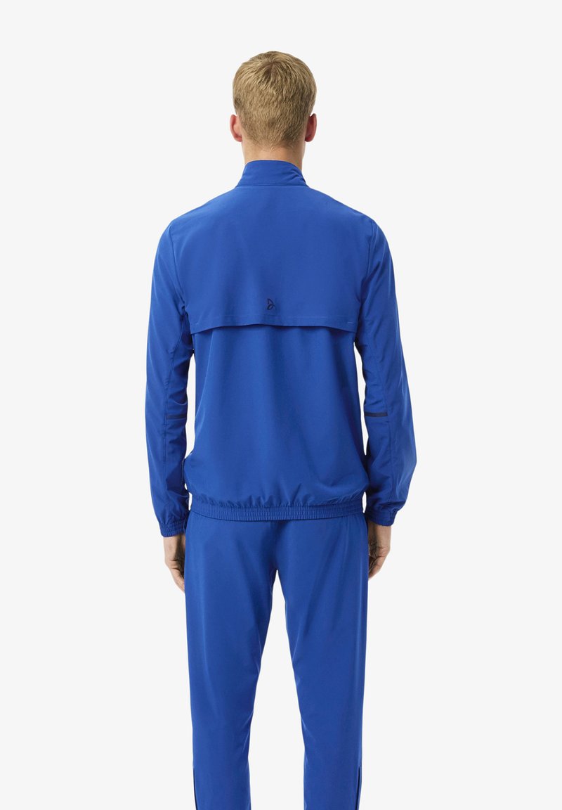 Mann mit kurzen blonden Haaren, der in einem blauen Trainingsanzug steht und von hinten vor einem schlichten weißen Hintergrund blickt.