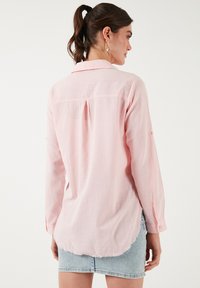 Light pink long-sleeve shirt with a collared neckline, a back pleat, and a curved hem. Made from textured fabric. Worn over a denim skirt.