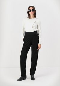 Pantalons noirs à jambes larges associés à un pull blanc. Le mannequin porte de grandes lunettes de soleil noires et des bottines. Tissu lisse et coupe décontractée.
