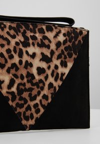 Pochette avec imprimé léopard et panneaux en daim noir dans un design géométrique, avec une lanière de poignet sur un fond uni.