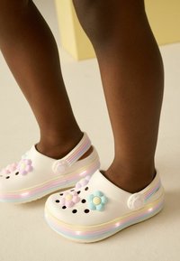 Gambe di un bambino che indossano zoccoli bianchi con suole a strisce pastello, decorazioni floreali e luci LED rosa su un pavimento chiaro.