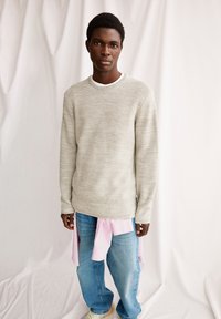 Hellgrauer gestrickter Pullover mit Rundhalsausschnitt, kombiniert mit blauen Jeans und einem hellrosa Hemd, das um die Taille gebunden ist.