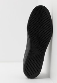 Semelle de chaussure noire avec des rainures diagonales et un logo crocodile embossé au centre, photographiée sur un fond blanc en coin.