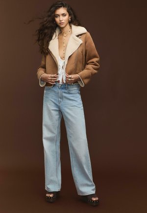 Veste en shearling marron avec doublure crème, top blanc avec franges, jeans larges en denim bleu clair et sandales à plateforme noires avec des clous.