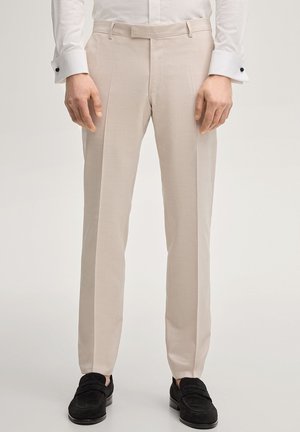 Pantaloni beige su misura con pieghe frontali indossati con una camicia bianca e mocassini neri su uno sfondo neutro.