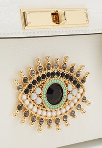 White leather handbag featuring a prominent gold-tone eye embellishment with black, white, and turquoise beads, creating a detailed pattern.