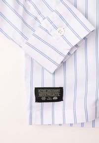 Λευκό πουκάμισο με ανοιχτό μπλε κάθετες ρίγες, με μανσέτα με κουμπιά και μαύρη ετικέτα με κείμενο και γραφικά στοιχεία κοντά στο τελείωμα.