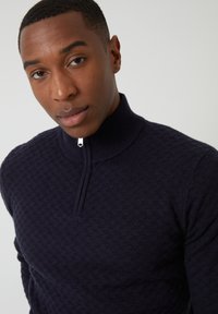 Granatowy sweter z dzianiny z teksturowanym wzorem, wysokim kołnierzem i zamkiem błyskawicznym do pół. Materiał wydaje się być miękki i dziany. Brak dodatkowych widocznych szczegółów.