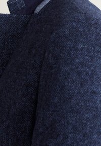 Giacca in lana texturizzata di colore navy con un colletto alto e dettagli di cucitura sottili. Il tessuto appare morbido con una finitura leggermente ruvida.