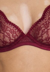 Soutien-gorge en dentelle bordeaux avec des bonnets triangulaires, des détails en dentelle complexes et une bande élastique lisse au bas. Pas de matériel visible.