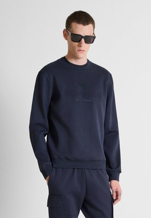 Man draagt donkerblauwe sweatshirt en bijpassende cargobroek, met zwarte rechthoekige zonnebril, staand tegen een effen lichte achtergrond.