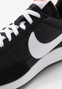 Baskets noires avec lacets blancs et un grand swoosh blanc Nike sur le côté, montrées en gros plan sur fond blanc.