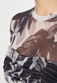 Gros plan sur un torse portant un haut transparent à manches longues avec un grand imprimé floral et un détail froncé en tissu noir et gris sur la poitrine.