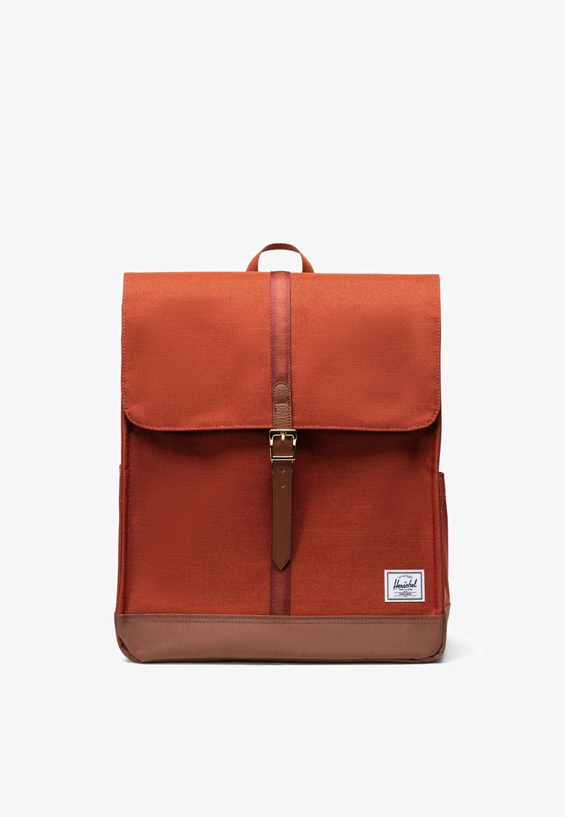 Mochila de color óxido con un tejido texturizado, un fondo de cuero marrón y un cierre de hebilla en la parte frontal. Cuenta con un asa superior y un parche con el logotipo.