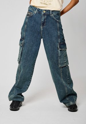 Person iført blå denim cargobukser med vide ben og store sidelommer samt sorte sneakers, stående foran en ensfarvet baggrund.