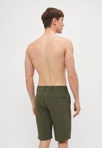 Olivenfargede shorts laget av lett stoff, med to baklommer og et enkelt logomerke på venstre lomme.