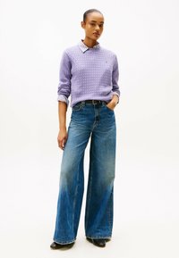 Fioletowy sweter z dzianiny z wysokim kołnierzem, w połączeniu z szerokimi niebieskimi dżinsami i czarnymi botkami do kostek, na tle gładkiego białego tła.
