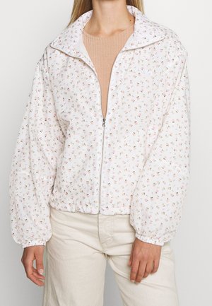 Femme portant une veste blanche zippée avec un petit imprimé floral et des poignets élastiques, associée à un pantalon beige et un haut côtelé beige.