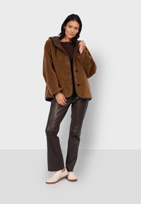 Braune Fleecejacke mit Kapuze, mit einem Reißverschluss vorne, drei Knöpfen und kontrastierenden schwarzen Nähten, kombiniert mit dunkelbraunen Kunstlederhosen und Sneakers.