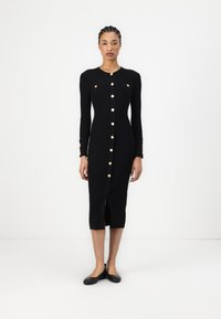 Robe noire à côtes avec des manches longues, fermetures à boutons dorés, fente à l'avant et deux poches poitrine. Accents cousus sur les manches.